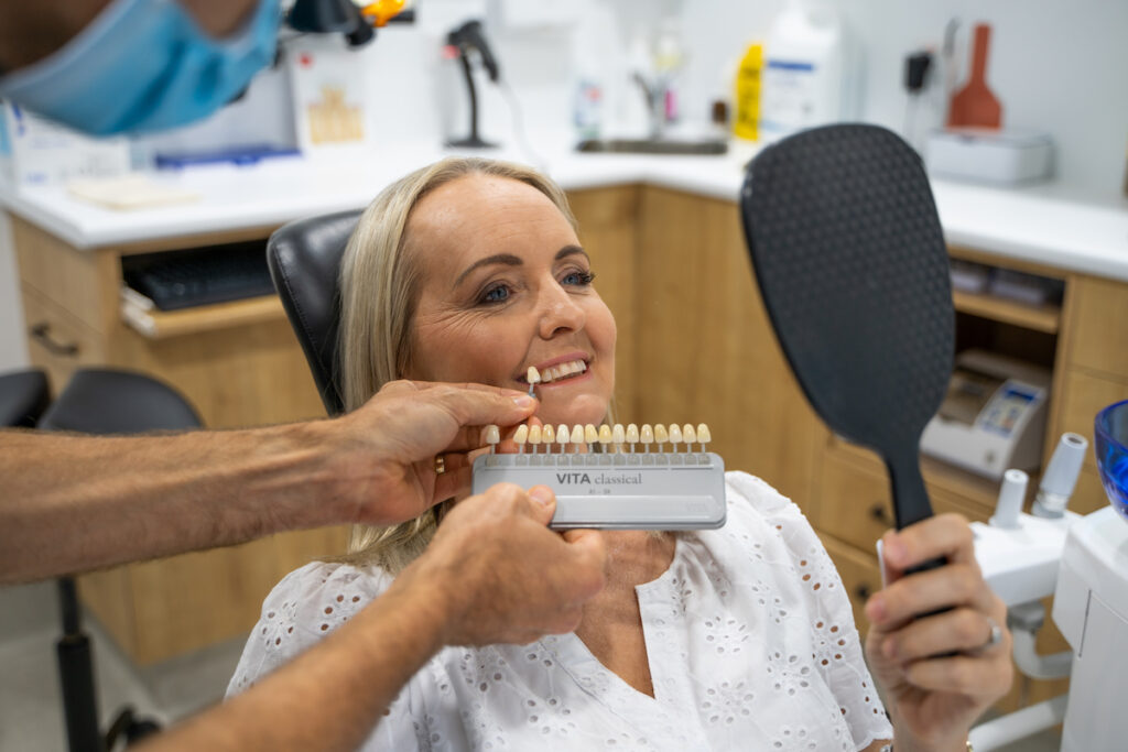 A patient looks on with a mirror as Dr Nick uses a shade guide to determine the colour of her natural teeth.