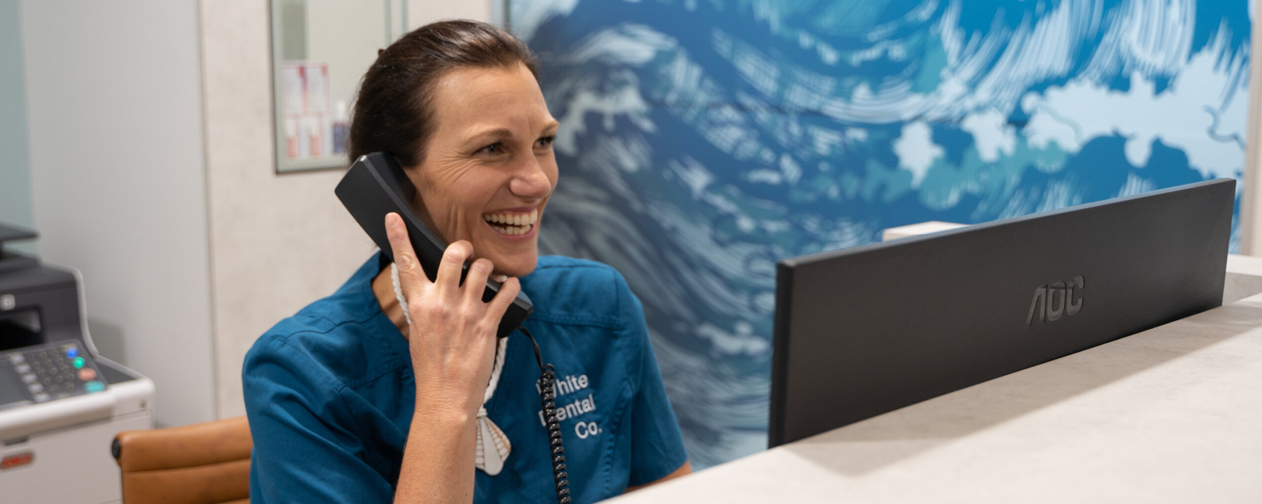 Dental Assistant Kelly sits at White Dental Co.'s reception desk, making an emergency dental appointment for a patient who is calling on the phone.