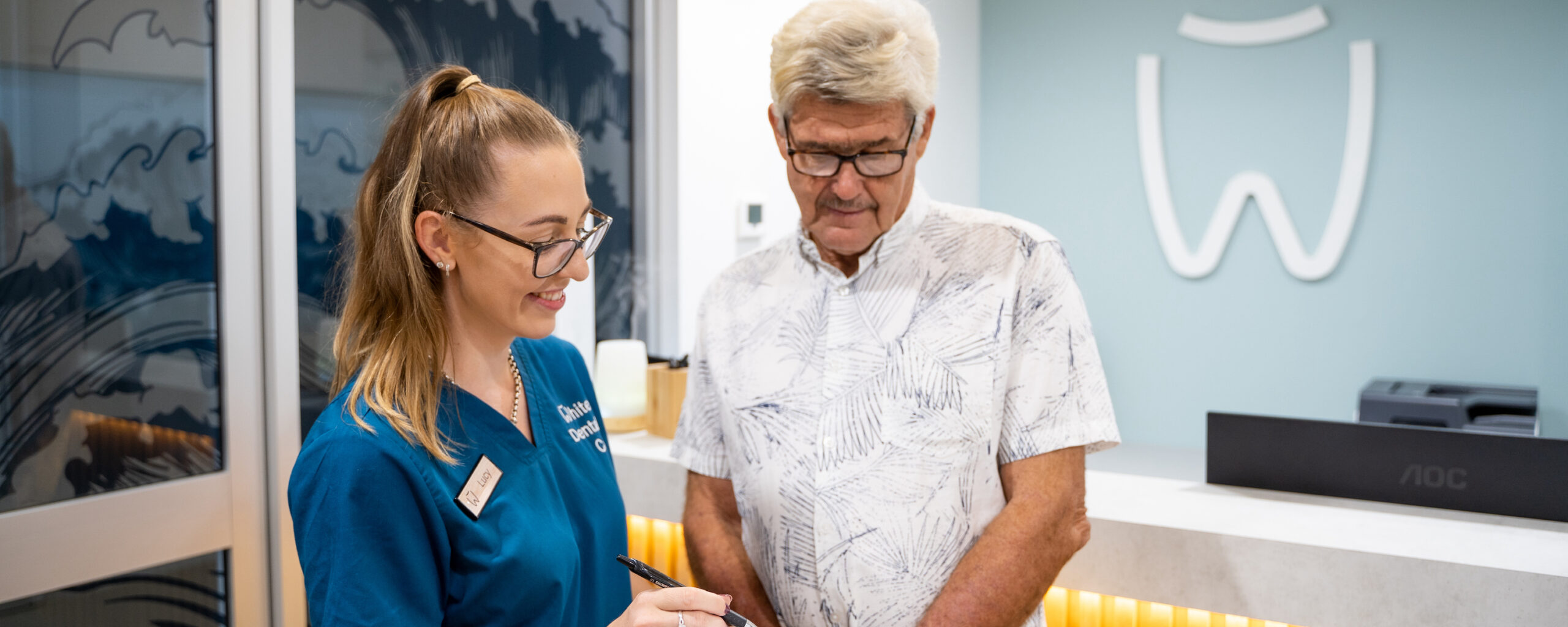 Dental assistant Lucy smiles as she assists a senior patient at the reception desk