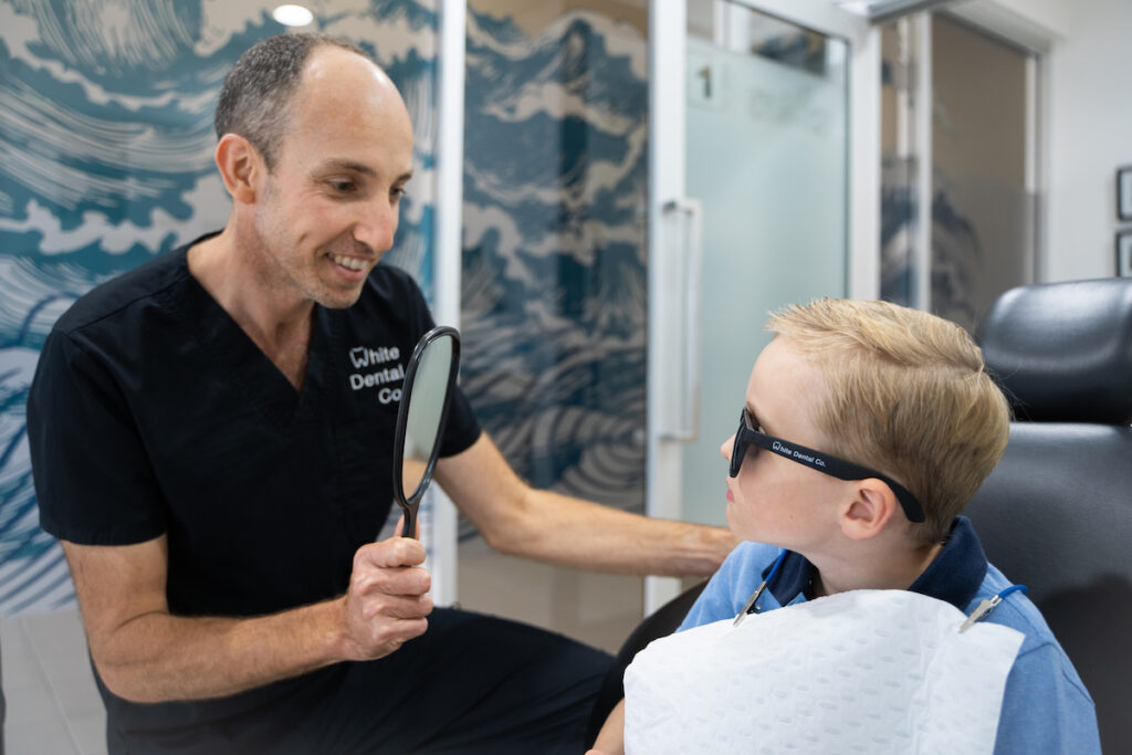 Dr Nick, dentist at white Dental Co., holds a mirror up for a young patient to look at their teeth at the end of their dental check-up appointment