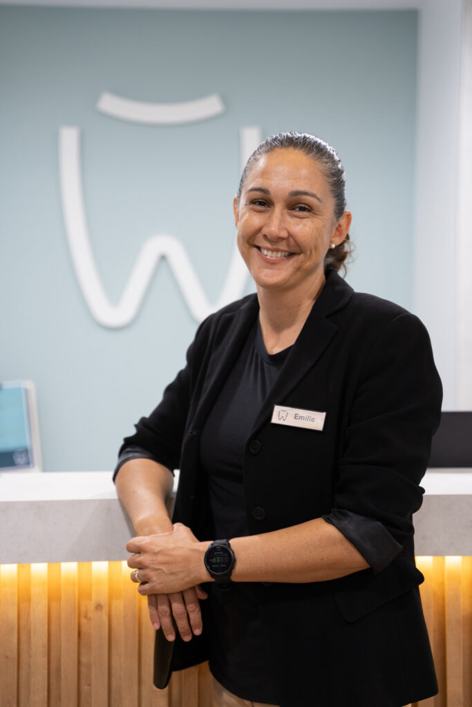 Emilie stands smiling in front of the reception desk. The White Dental Co. logo is visible on the wall behind her.